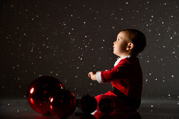 Little toddler boy in santa costume is sitting on the floor at christmas tree balls looking up under the snow