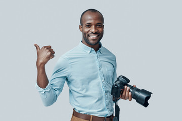 Happy young African man using digital camera and smiling while standing against grey background