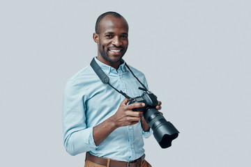 Happy young African man using digital camera and smiling while standing against grey background