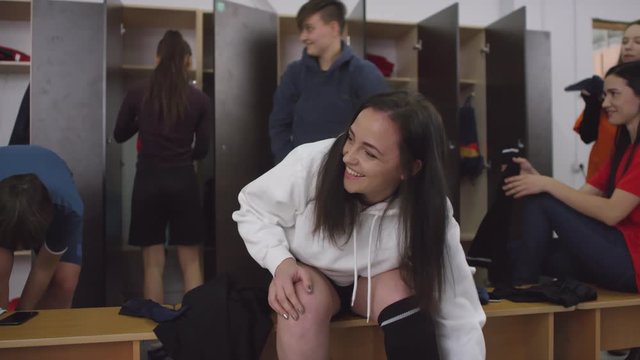 Young beautiful sportswoman sitting on bench in locker room, smiling and chatting with female teammates while changing her clothes before soccer workout