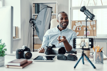 Happy young African man in shirt showing digital camera and telling something while making social media video