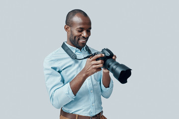 Happy young African man using digital camera and smiling while standing against grey background