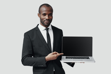 Confident young African man in formalwear pointing copy space on laptop and smiling while standing against grey background