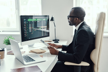 Concentrated young African man in formalwear using modern technologies while working in the office