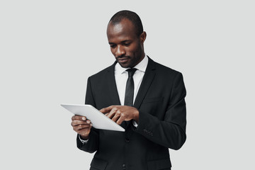 Concentrated young African man in formalwear working using digital tablet while standing against grey background