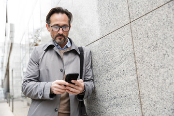 Image of serious businessman using cellphone while leaning on wall