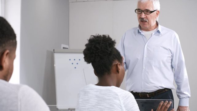 Over-shoulder tilting medium shot of strict senior Caucasian male professor speaking in front of multiethnic students, sitting at desks and working on touchscreen AR tablets. Template for VR graphics.