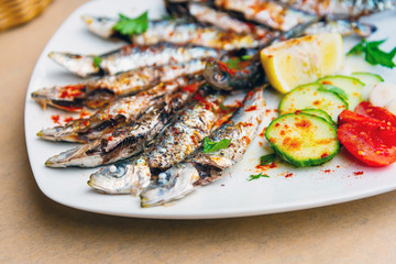 Fresh Grilled sardines served with fresh tomatoes, cucumbers slices and lemon on white plate for mediterranean healthy meal