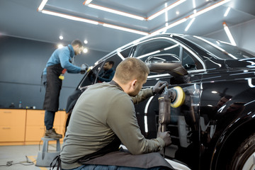 Two workers polishing vehicle body with special grinder and wax from scratches at the car service...