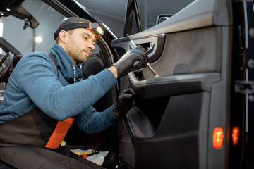 Car service worker in uniform provides a professional vehicle interior cleaning, wiping door panel with a brush at the car service station