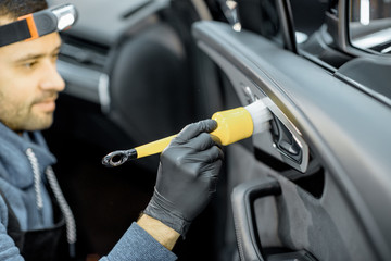 Car service worker in uniform provides a professional vehicle interior cleaning, wiping door panel with a brush at the car service station