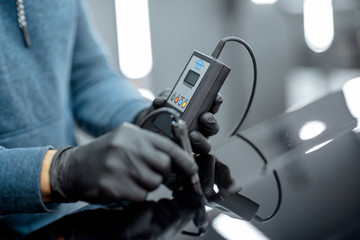 Inspector measuring paint cover thickness of the car body at the vehicle service box, close-up view on hands and professional thickness meter