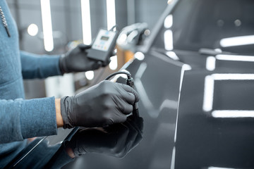 Inspector measuring paint cover thickness of the car body at the vehicle service box, close-up view on hands and professional thickness meter