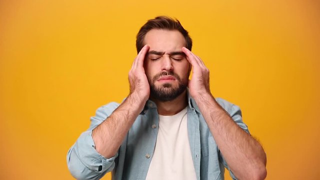 Sad upset bearded young guy 20s in denim shirt white t-shirt isolated over yellow background in studio. People sincere emotions, lifestyle concept. Looks at camera, rubs temples with hands, headache.