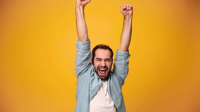Handsome bearded young guy 20s in denim shirt white t-shirt isolated over yellow background in studio. People sincere emotions, lifestyle concept. Looking at the camera doing winner gesture, say Yes