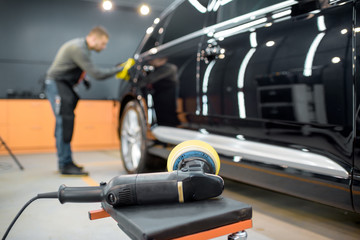 Car service worker wiping vehicle body with microfiber, examining glossy coating after the polishing procedure. Grinder on the foreground