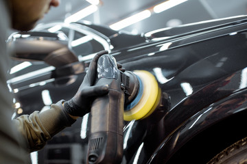 Worker polishing vehicle body with special grinder and wax from scratches at the car service station. Professional car detailing and maintenance concept