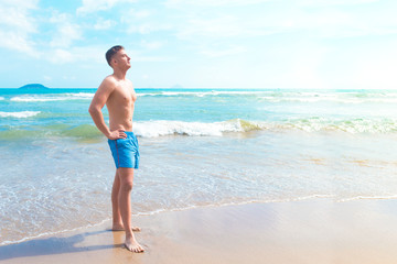 Full length portrait of handsome beautiful fit slim athletic guy in swimming trunks on the sand beach at blue sea. Young man enjoying tropical vacation, summer sunny weather, sunbathing, swimming.