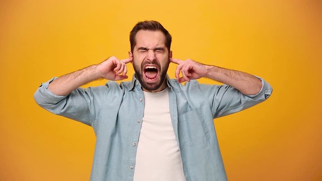 Angry bearded young guy 20s in denim shirt white t-shirt isolated over yellow background in studio. People sincere emotions, lifestyle concept. Looking at the camera. Covering ears with fingers scream