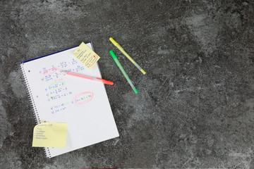 mathematics book with calculation tasks, with orange, yellow and green pencil, shopping list and to do list post it on a grey marble table