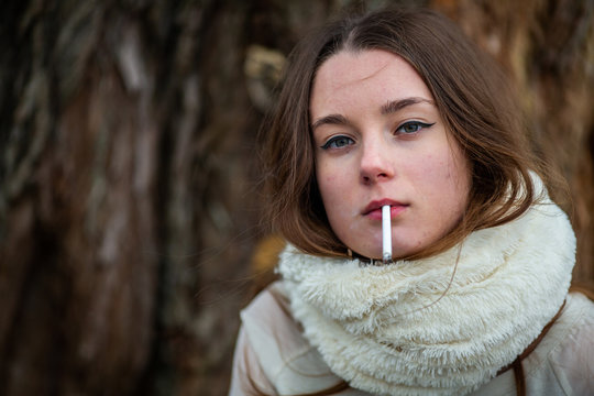 Smoking teenager in real life. Young beautiful white teeage girl in scarf smokes an cigarette near the tree on the street in the autumn. Deadly bad habit.