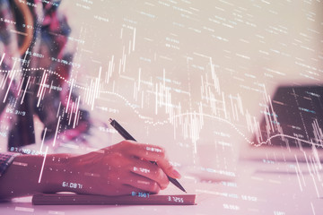 A woman hands writing information about stock market in notepad. Forex chart holograms in front. Concept of research. Double exposure