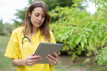 Fototapeta premium Mujer enfermera joven vestida con uniforme amarillo y estetoscopio colgado del cuello observando con atención la tablet que sostien con ambas manos en entorno de naturaleza