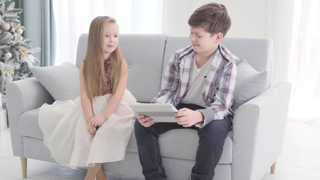 Charming girl in beautiful dress touching boy's shoulder and waving. Brunette guy making displeased face and keeping watching movie on tablet. Offended lady crossing hands.