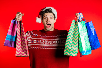 Happy young man in Santa hat with colorful paper bags after shopping isolated on red studio background. Winter sale, purchases, spending money on gifts concept.