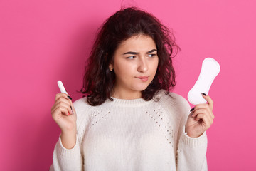 Indoor shot of young brunette woman holding two intimate products, choosing between tampon and pad during menses, thoughtful lady wearing white sweater. Women health care and hygiene concept. © sementsova321