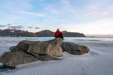 Man in red jacket sits on Ramberg beach in Lofoten island in Norway during sunset. Houses and mountains in the background. Travelocity and traveling concept.