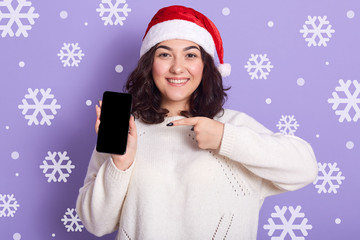 Closeup portrait of smiling magnetic cute dark haired female pointing at smartphone with forefinger, holding mobile phone in one hand, looking directly at camera. Present alternatives concept.