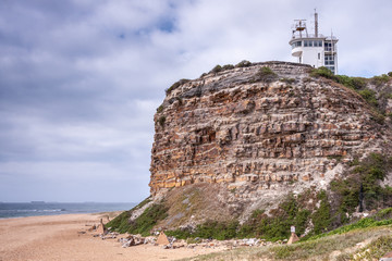 Newcastle, Australia - December 10, 2009: Nobbys Lighthouse buildings on brown-gray cliffl under blue cloudscape. Sandy beach and blue-gray South Pacific Ocean water. Bulk ship on horizon.