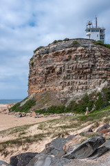 Fototapeta na wymiar Newcastle, Australia - December 10, 2009: Closeup of Nobbys Lighthouse buildings on brown-gray cliffl under blue cloudscape. Sandy beach and blue-gray South Pacific Ocean water. 