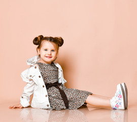 Kid girl with funny buns in leopard print dress, denim jacket is sitting on the floor sweet smiling
