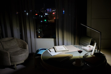 Place of work at the room with working table, armchair and large window with night city view.