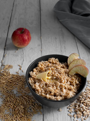 Healthy breakfast. Oatmeal with apple slices and butter. Black cup with porridge on a white wooden background. Oat grains are scattered on the table. Close-up.