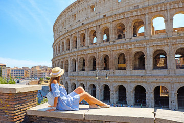 Travel woman in romantic dress and hat sitting and looking on Coliseum, Rome, Italy. Beautiful tourist girl with backpack near Colosseum. Young woman enjoy summer Italian vacation in Europe. - obrazy, fototapety, plakaty