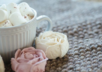A cup of cocoa or chocolate next to a rose-shaped marshmallow. Warm cozy square background.