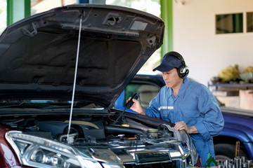 Asian automotive mechanic with blue uniform use electronic tools to check the car in part of engine near hood in the garage.
