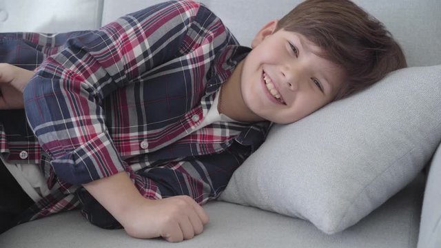 Portrait of smiling Caucasian brunette boy with grey eyes lying on sofa at home. Happy child resting indoors on weekends. Leisure, childhood, lifestyle.