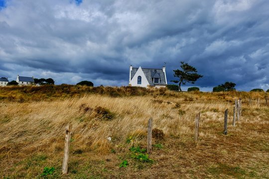 Panorama rurale con un cottage tra l&rsquo;erba secca. Un cielo drammatico avvolge la scena. Panorama bretone