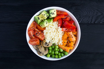 Vegan poke bowl with couscous  and vegetables in the white bowl in the center of the black wooden background. Top view. Copy space. Closeup.