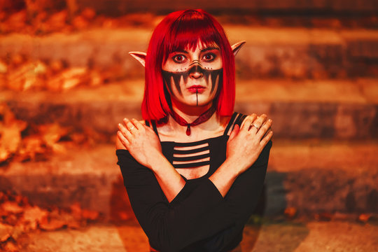 Girl cosplay in the image of a dark elf in a red wig portrait close-up. A woman with red lenses in her eyes and red hair in an elf costume with false ears and makeup. Dark elf outfit for Halloween.
