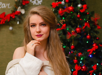 Portrait of a girl with bright red lips, blond long hair against the background of a New Year tree. Young girl in a white warm coat, shoulders are visible. Holidays. Merry Christmas.