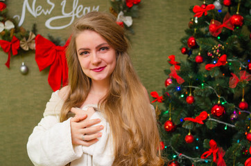 Portrait of a girl with bright red lips, blond long hair against the background of a New Year tree. Young girl in a white warm coat, shoulders are visible. Holidays. Merry Christmas.