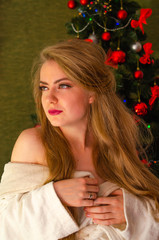 Portrait of a girl with bright red lips, blond long hair against the background of a New Year tree. Young girl in a white warm coat, shoulders are visible. Holidays. Merry Christmas.