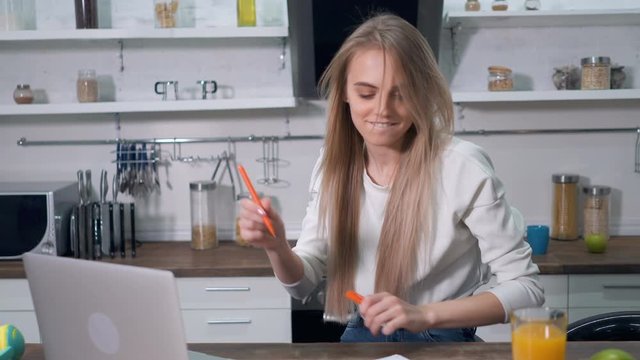 Blonde woman working with laptop at the kitchen. Sucess emotion, writing notes in the paper notebook. Handheld shot.