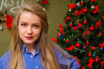 Portrait of a girl with bright red lips, blond long hair against the background of a New Year tree. Young girl in a blue men's shirt. Holidays. Merry Christmas. funny Face.