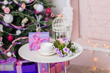 White table with a gift in a box, flowers, a cage and a cup on a saucer against the background of a Christmas tree and fireplace.